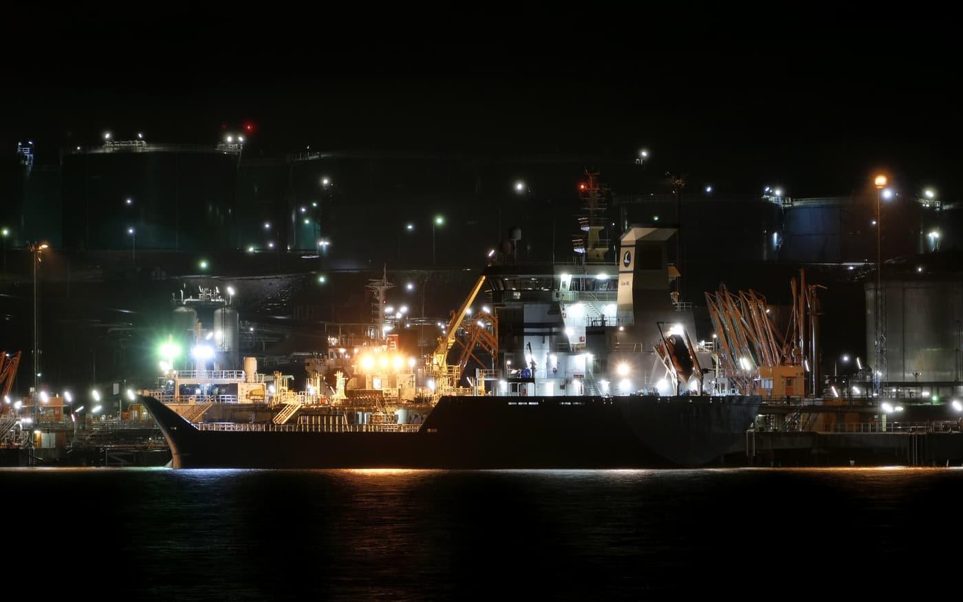 A products tanker berthed at an oil terminal at night, tank farm lit behind it.